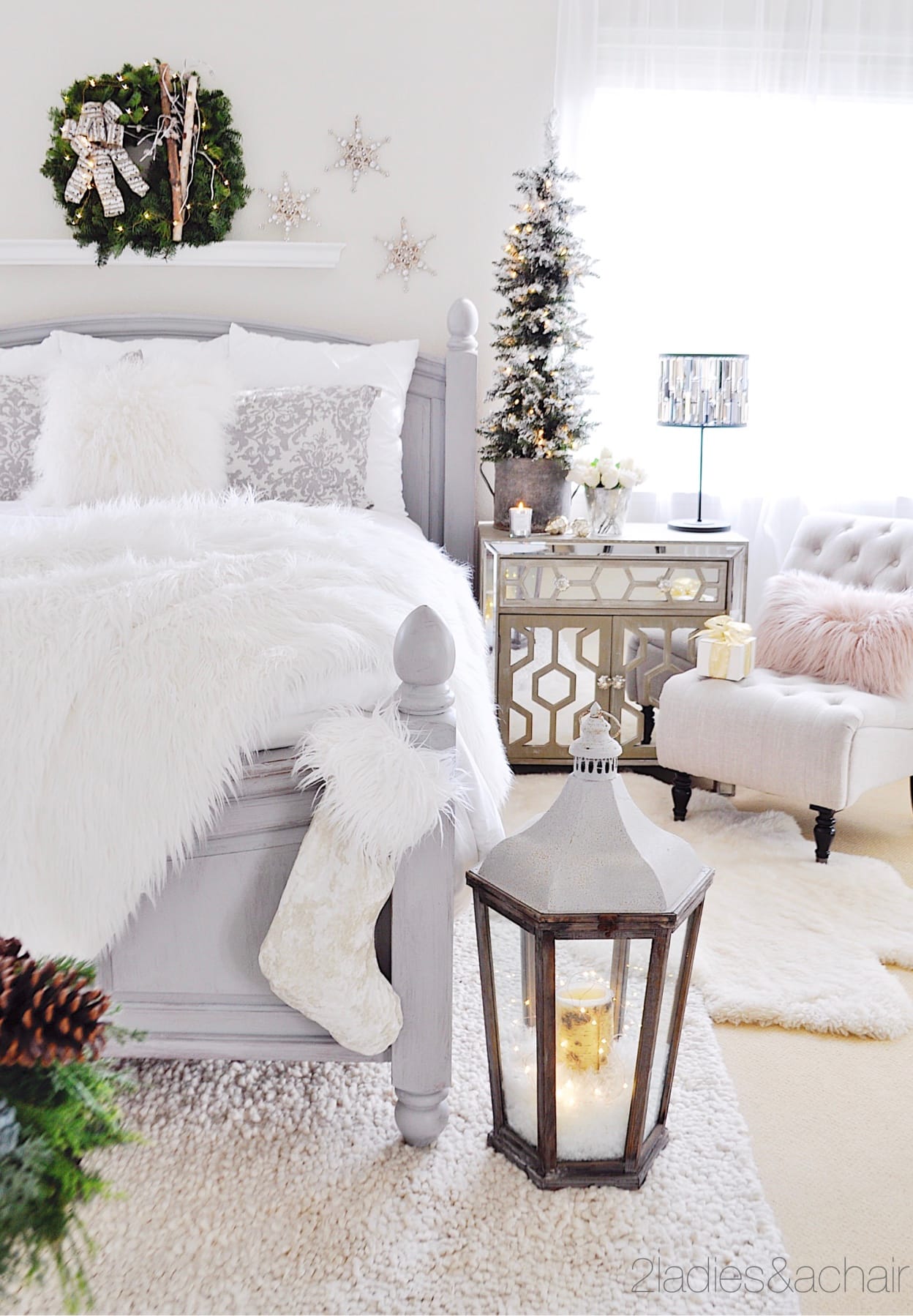 A flocked tree sits on a mirrored nighstand next to a bed with a plush white bedspread with Christmas decor scattered around the room
