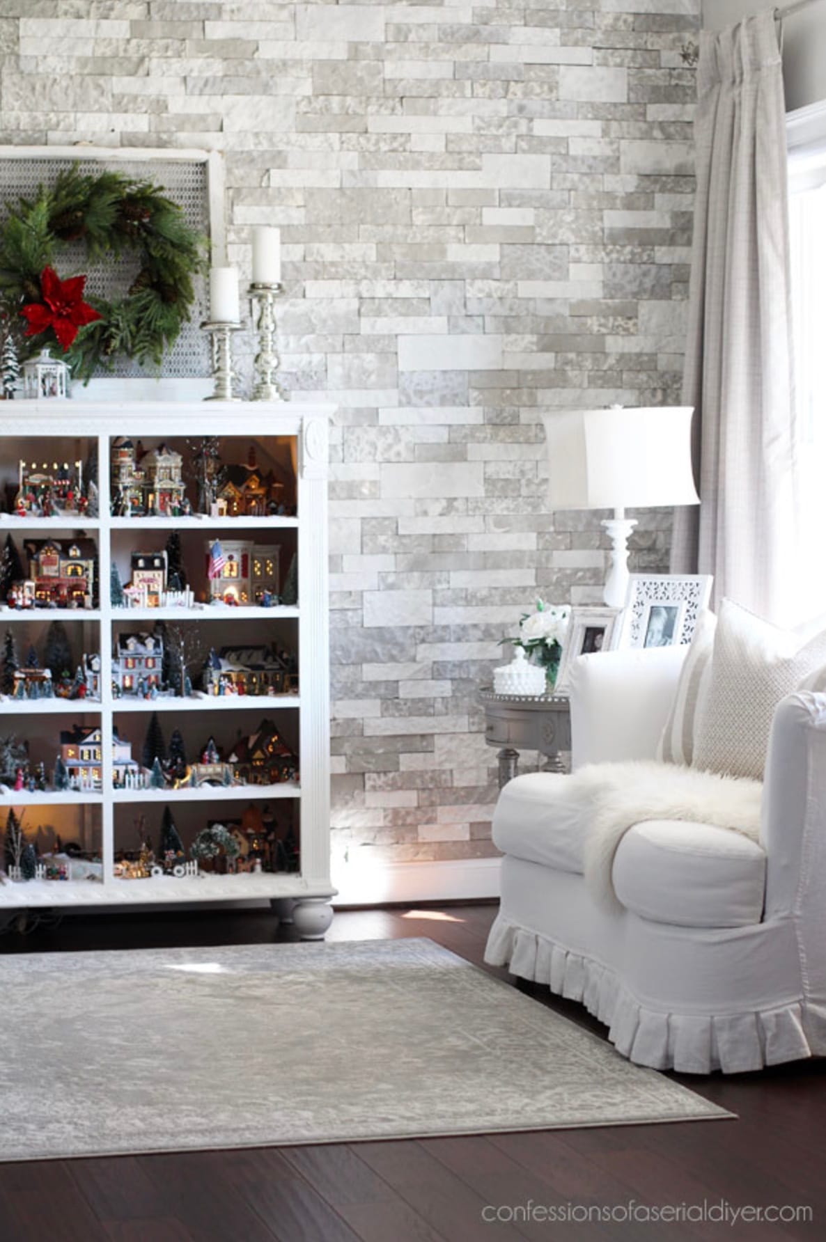 Christmas village displayed inside an empty chest of drawers with a wreath hanging above it in a white and gray living room