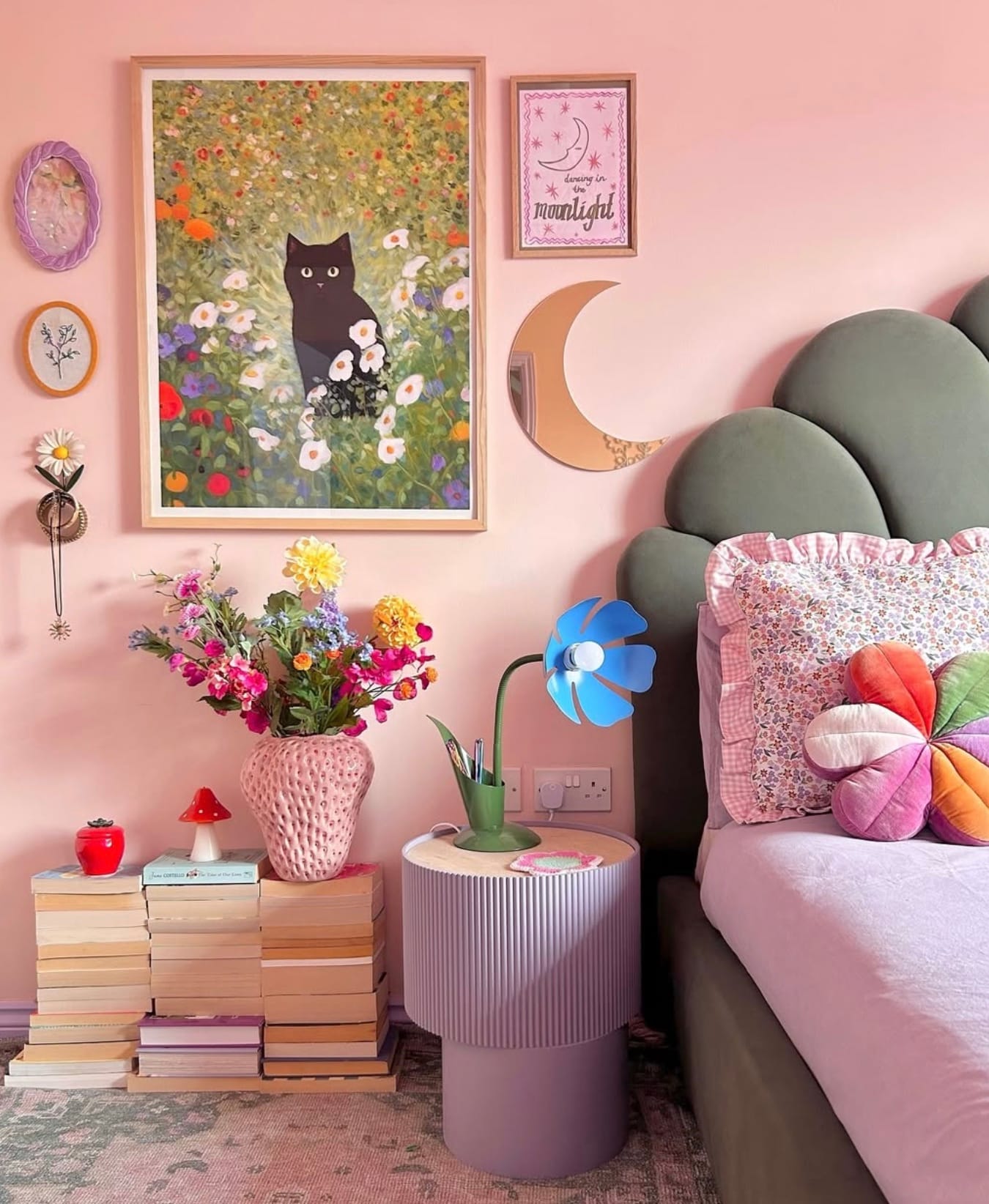 A light pink bedroom with a plush green headboard, cozy pillows, artwork, book stacks