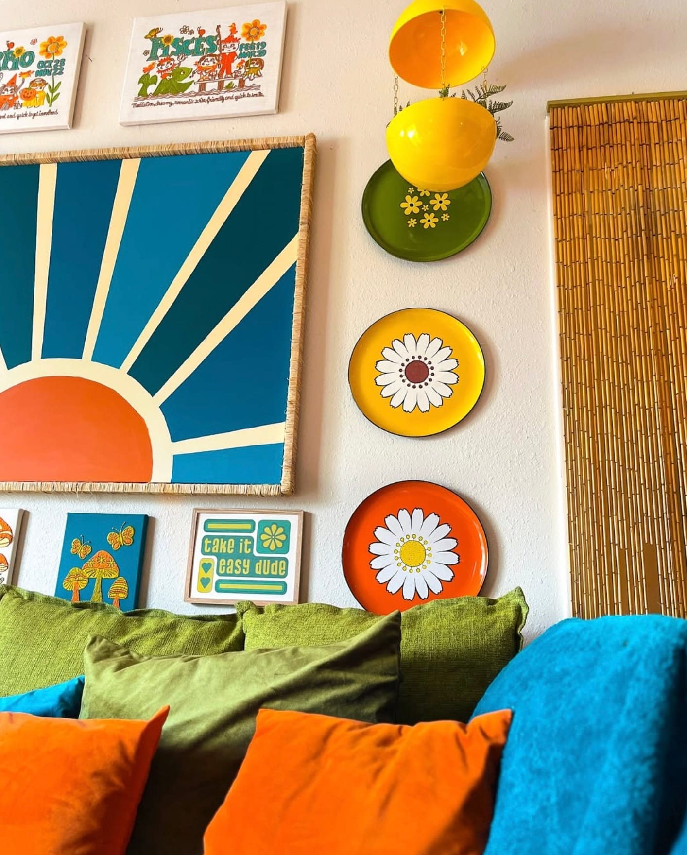 Colorful vintage plates are displayed next to artwork on the wall above a blue couch with green and orange cushions