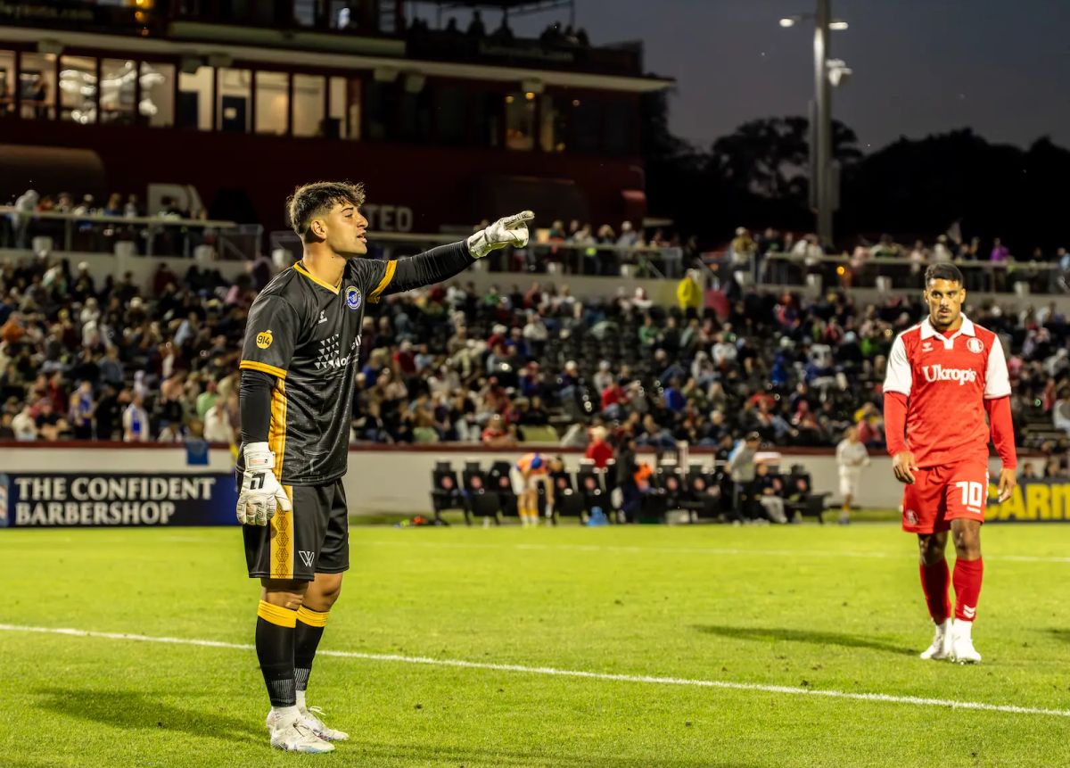 Westchester SC goalkeeper Luca Marinelli points to action off camera with a backdrop of a full stadium in Richmond as a Richmond player appears to walk towards the camera in the background
