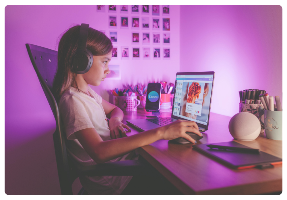 Young girl wearing headphones sits at a desk using a stylus and computer to design graphics, surrounded by pink and purple ambient lighting.