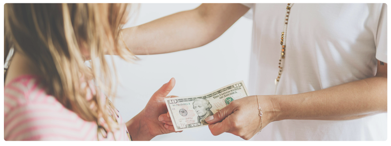Close-up of a child receiving a dollar bill from an adult, illustrating hands-on learning about saving, spending, and sharing money.