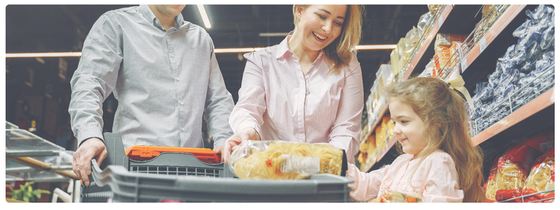 A parent and child grocery shopping together, with the child helping at checkout — modeling hands-on learning through everyday money decisions.