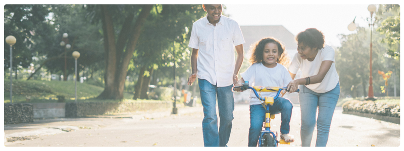 Parents helping their child learn to ride a bike in a park, illustrating resilience, encouragement, and growth through supported challenges.