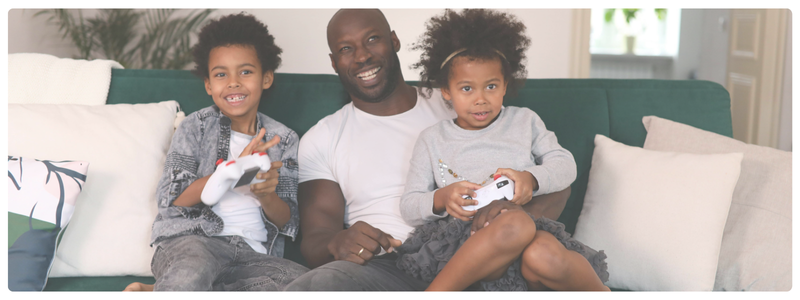 A smiling parent sitting on the couch with two kids playing video games, illustrating positive connection and support in a growth mindset environment.