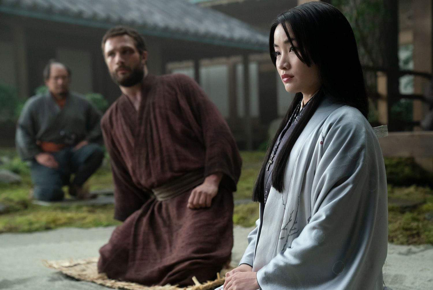 Cosmo Jarvis and Anna Sawai, along with a retainer, kneel in a courtyard in the Hulu mini-series Shogun