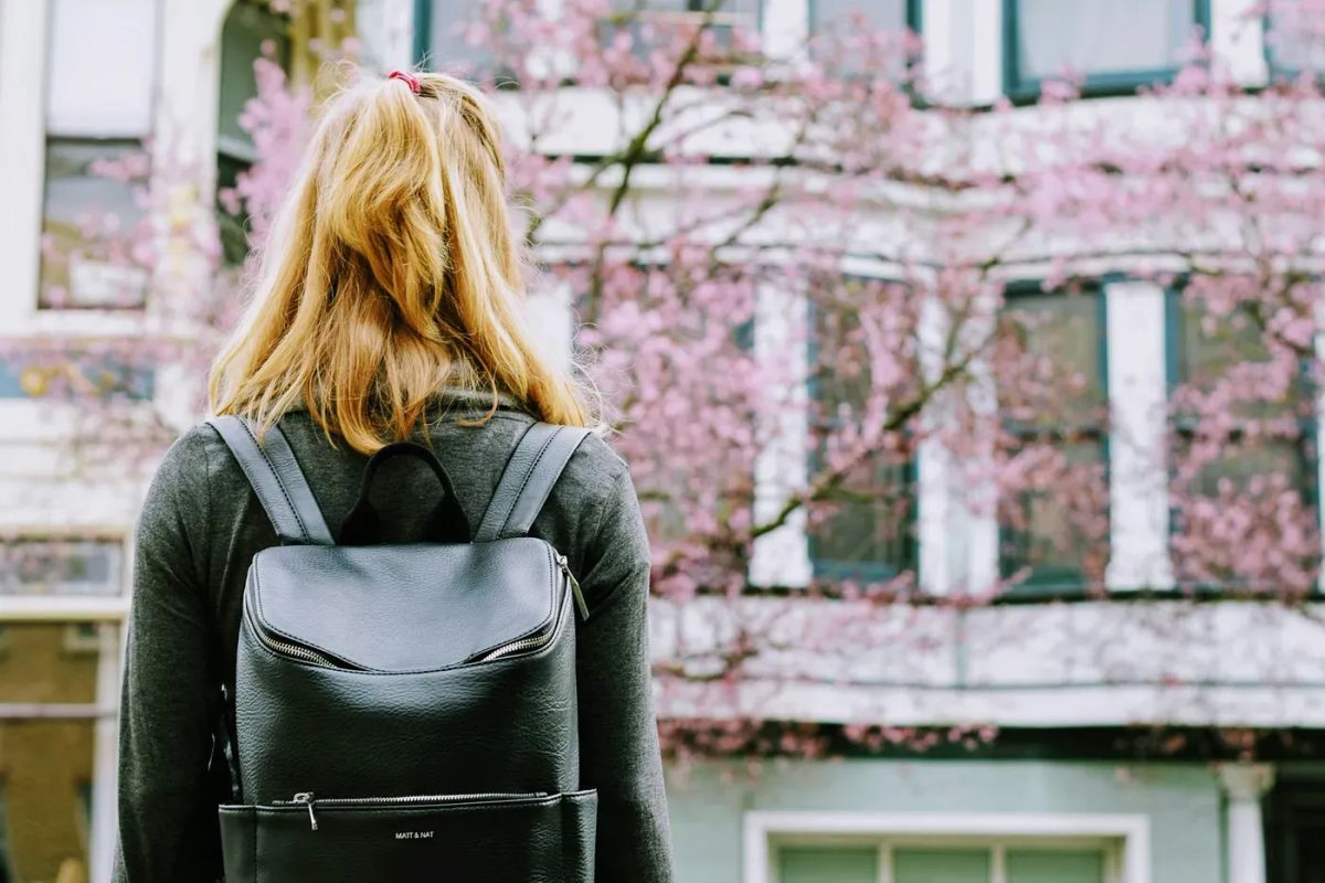 A blonde woman wearing a dark sweater and a leather backpack stands, facing away from the camera, looking toward an out-of-focus building and blooming tree with pink blossoms.