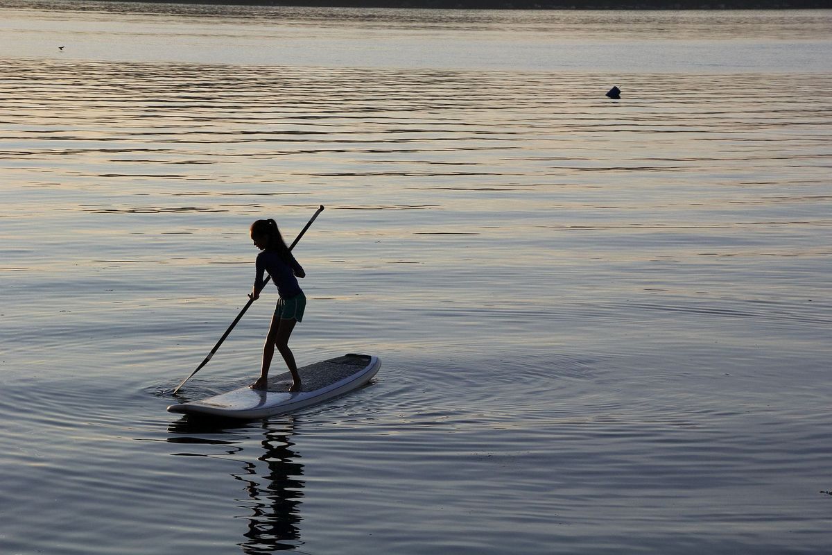 A young woman stands with an oar on a paddle board. She is surrounded by calm water reflecting the sunset.