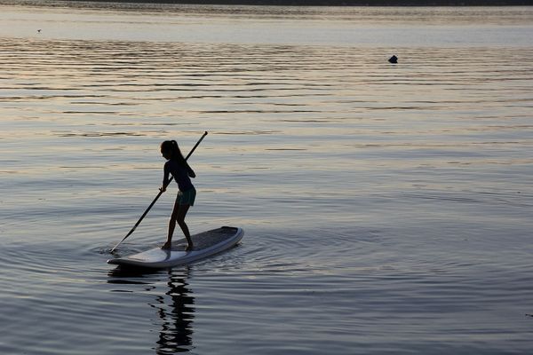A young woman stands with an oar on a paddle board. She is surrounded by calm water reflecting the sunset.