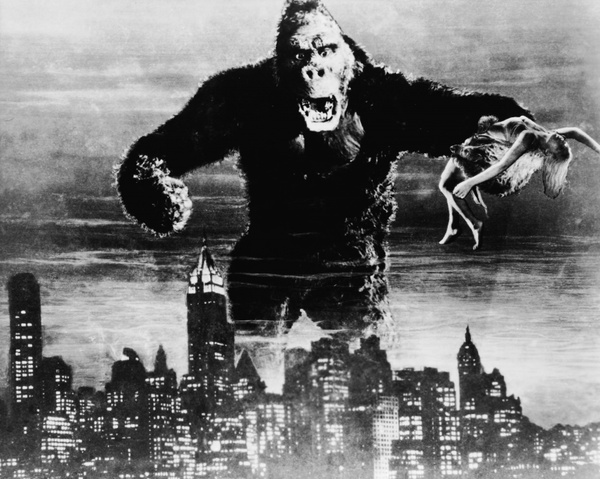 A gigantic, angy ape holds a woman on one had, towering over a brightly-lit city.
