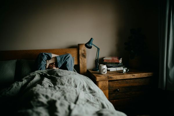 Person laying in bed next to a nightstand full of books, a lamp and a mug