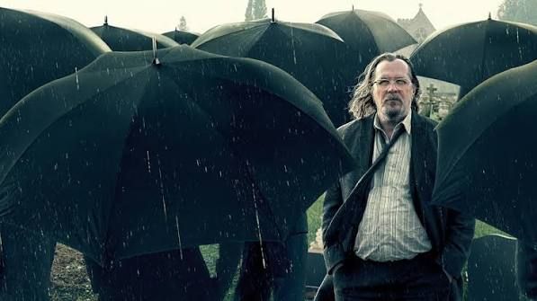 Gary Oldman, as the Slow Horses character Jackson Lamb, surrounded by black umbrellas on a rainy London day.