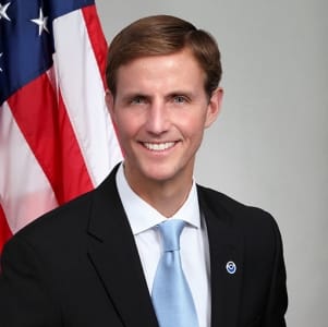 Portrait of Tim Gallaudet in a dark suit and light blue tie, standing before a United States flag.
