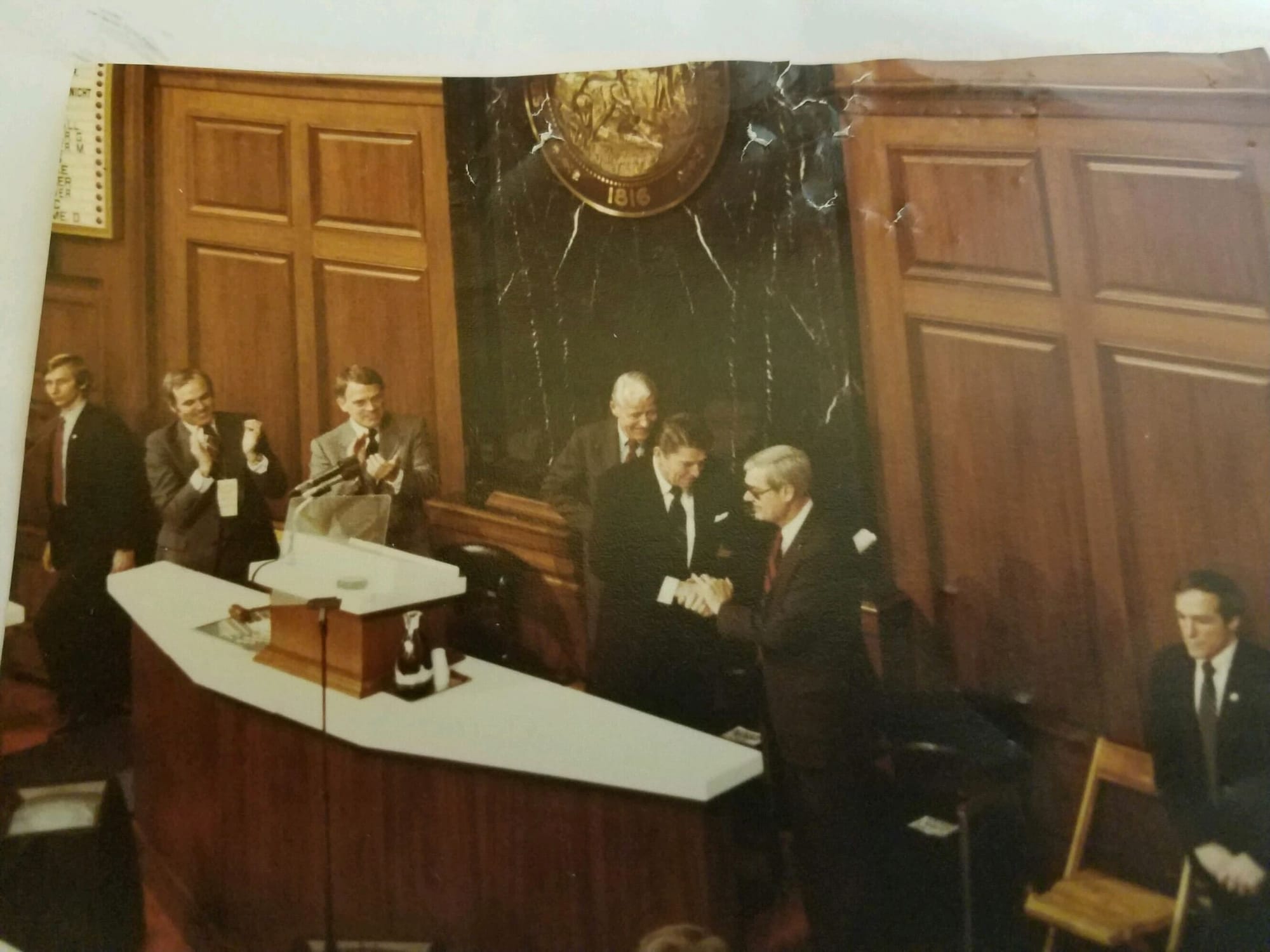 President Ronald Reagan shaking hands at the rostrum of a legislative chamber as those around him applaud, beneath a state seal dated 1816.