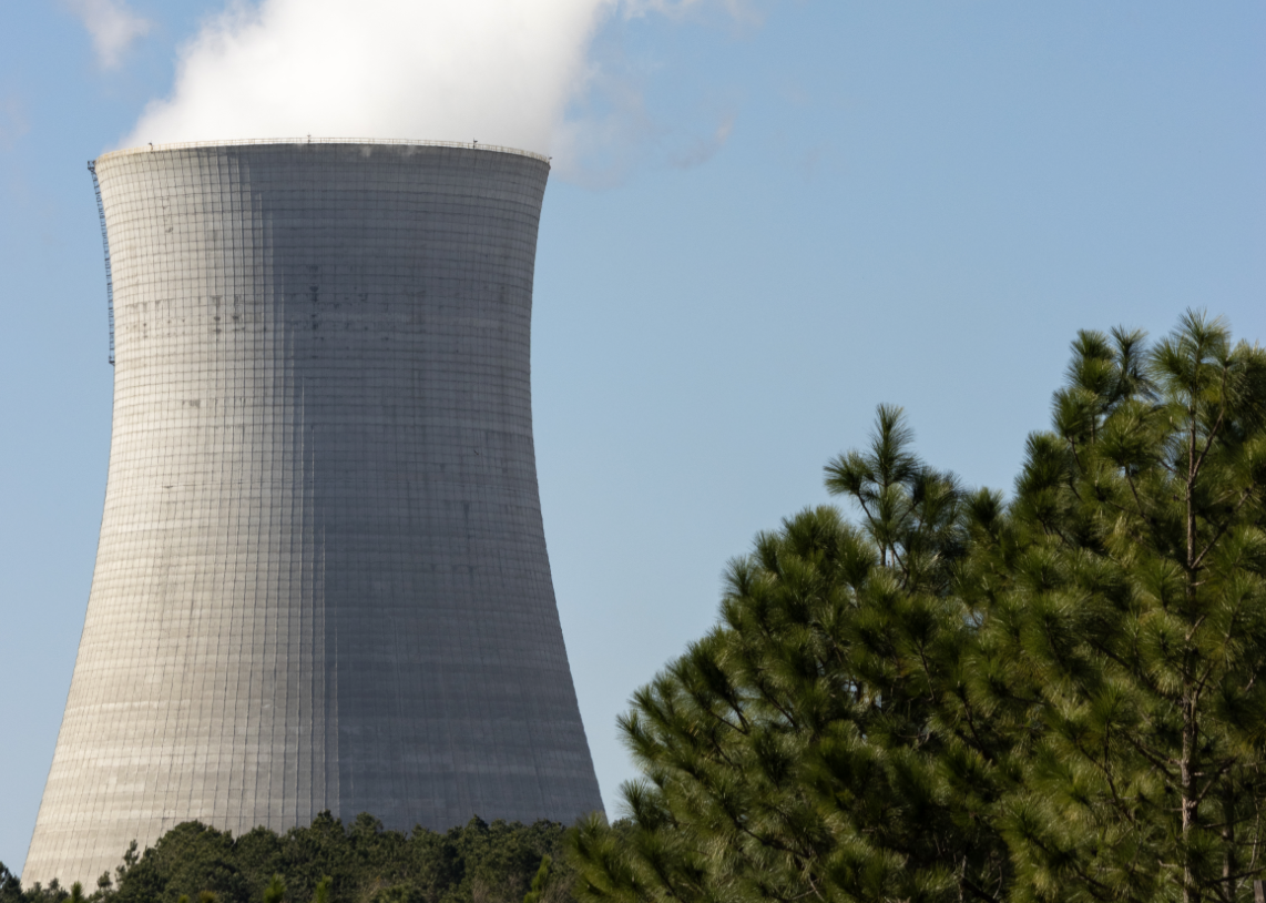 A concrete cooling tower at Plant Vogtle, steam rising from its rim, seen past a stand of Georgia pines.