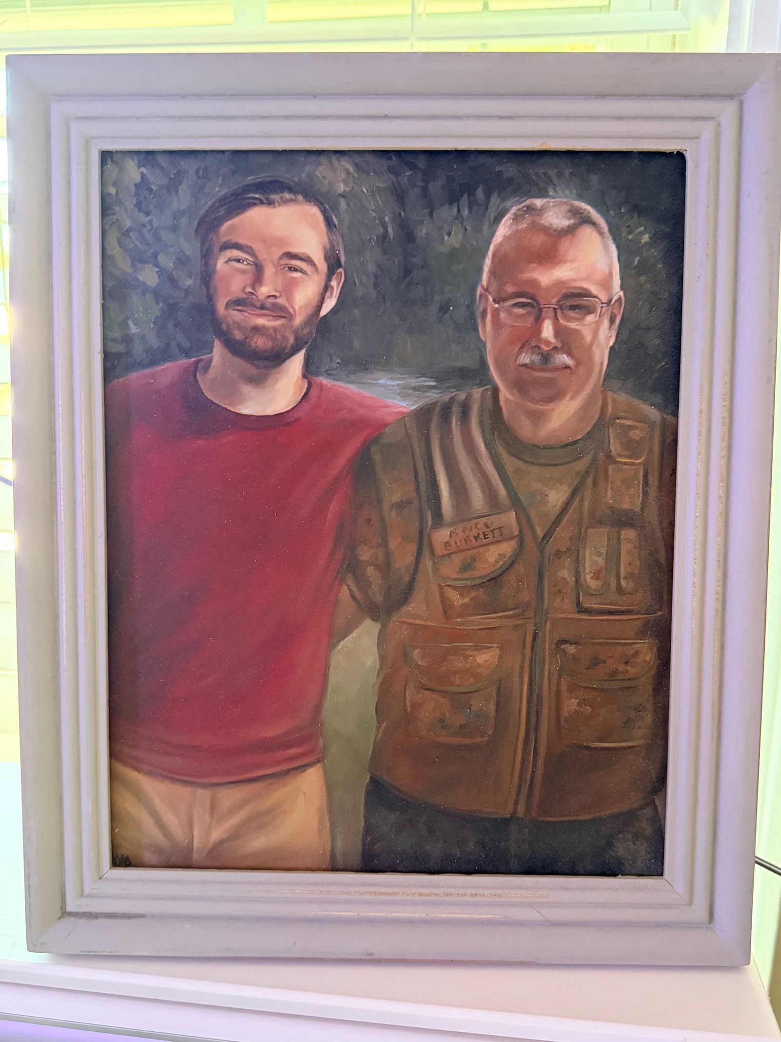 An oil portrait in a pale wooden frame: a bearded younger man in a red shirt with his arm around an older man in glasses wearing a tan multi-pocket vest with a Burkett name patch.