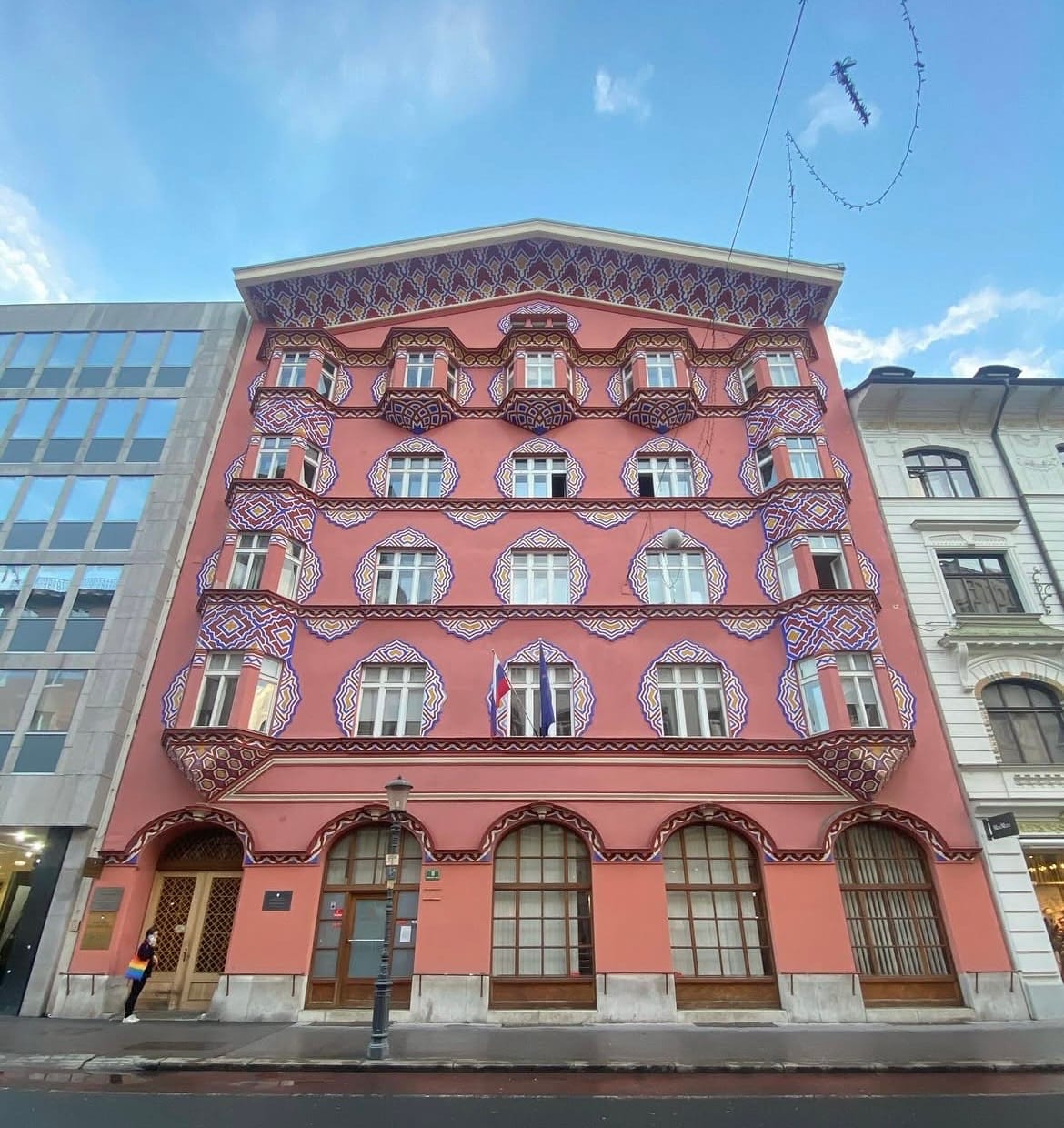Image of the facade of the Cooperative Business Bank Building in Ljubljana, Slovenia. It is a five-storey building. Its facade is reddish-orange, with windows and doors lined with red, yellow and blue geometric patterns