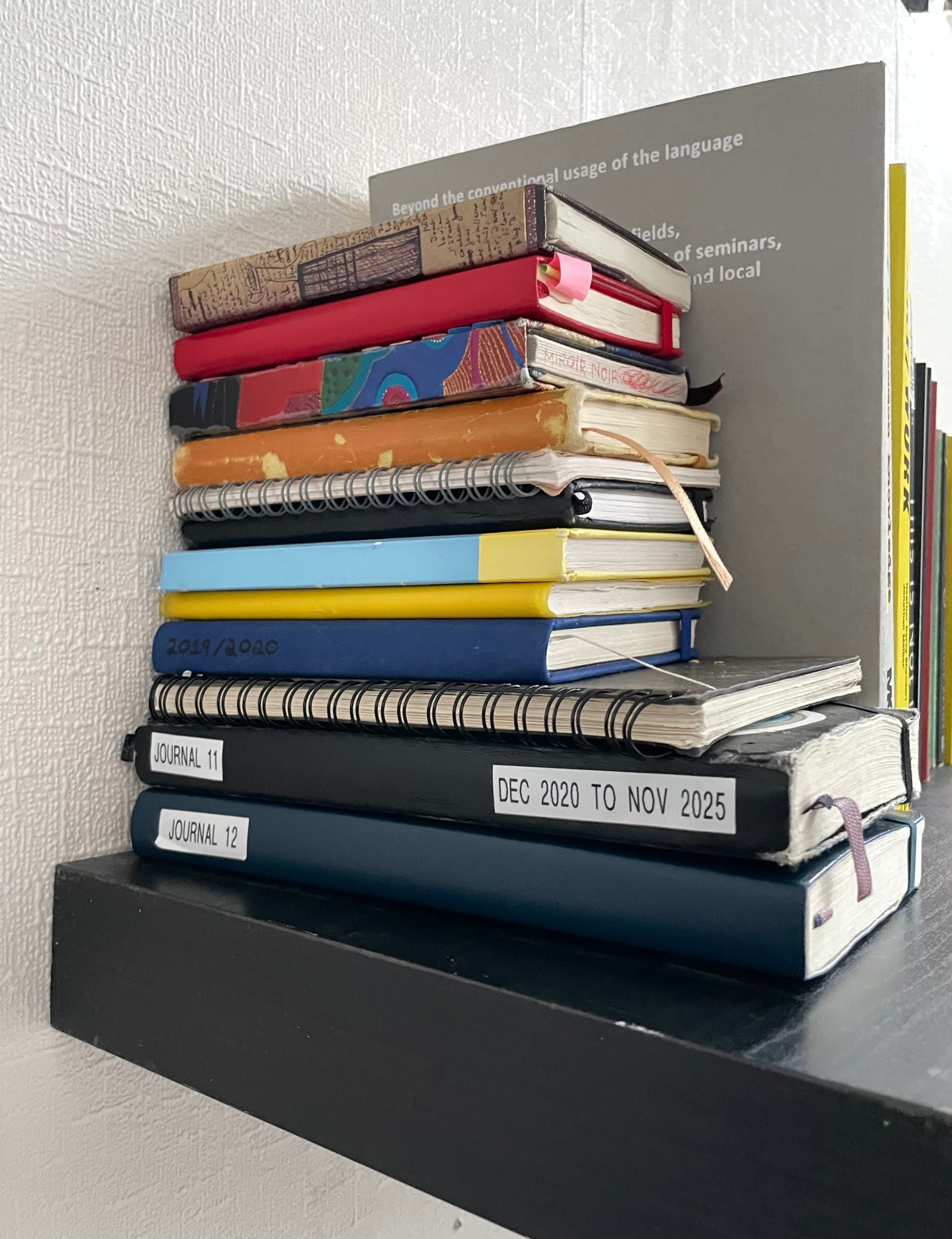 Image of a stack of my journals on a shelf. They are ordered from smallest on the top to largest on the bottom. One journal's spine has handwritten text on it that reads "2019/2020". Another spine reads "JOURNAL 11" and "DEC 2020 TO NOV 2025". Another spine reads "JOURNAL 12".
