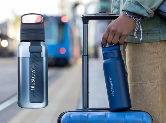 Traveler holding a water filter water bottle beside rolling luggage at a train station, showing portable hydration for trips and outdoor.