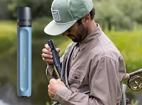 Fly fisherman using a water filter straw by a river, demonstrating portable water purification outdoors.