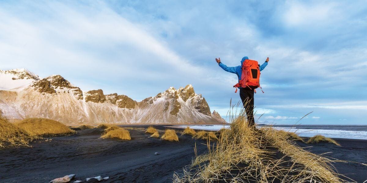Hiker with backpack standing on black sand beach, facing dramatic coastal mountains in Iceland.
