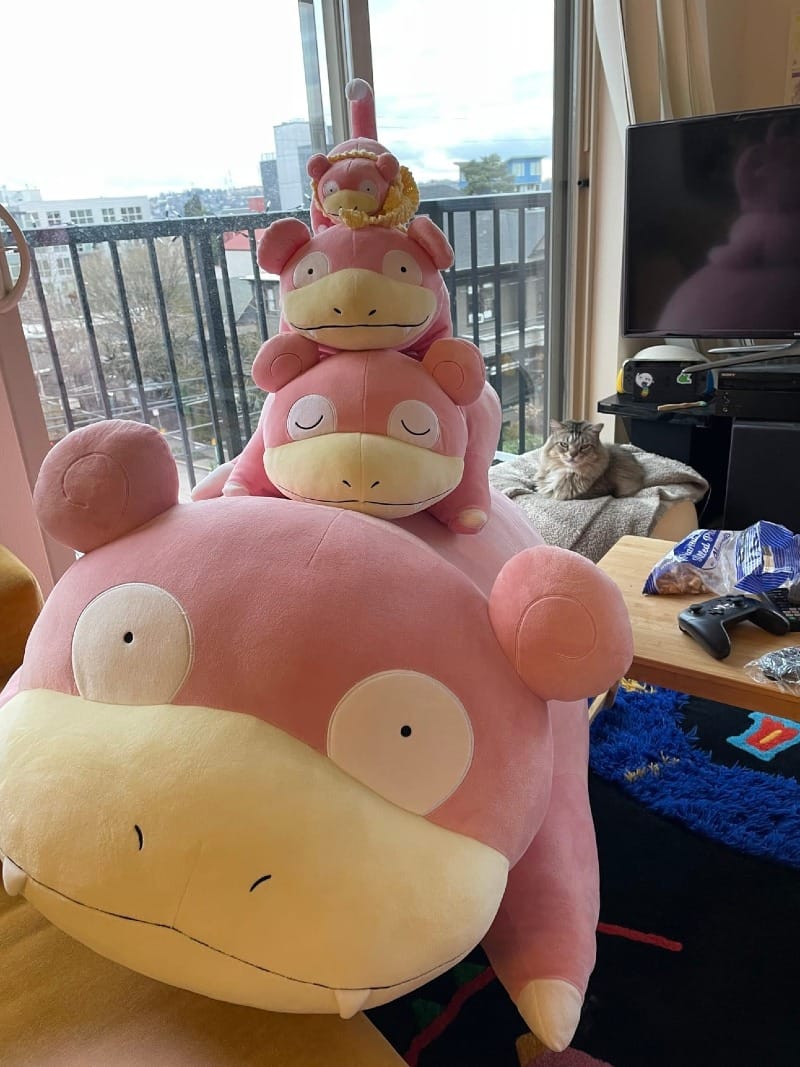 A tower of large pink plushies in a living room, with a cat sitting in the background.