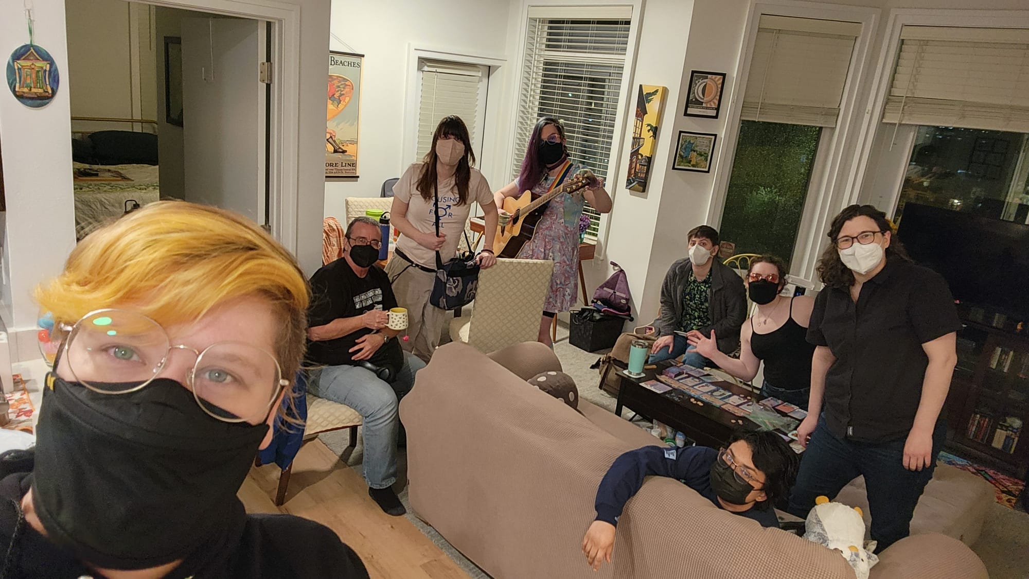 a group of folks ranging in hair length and ages, all wearing masks, lounging around a living room