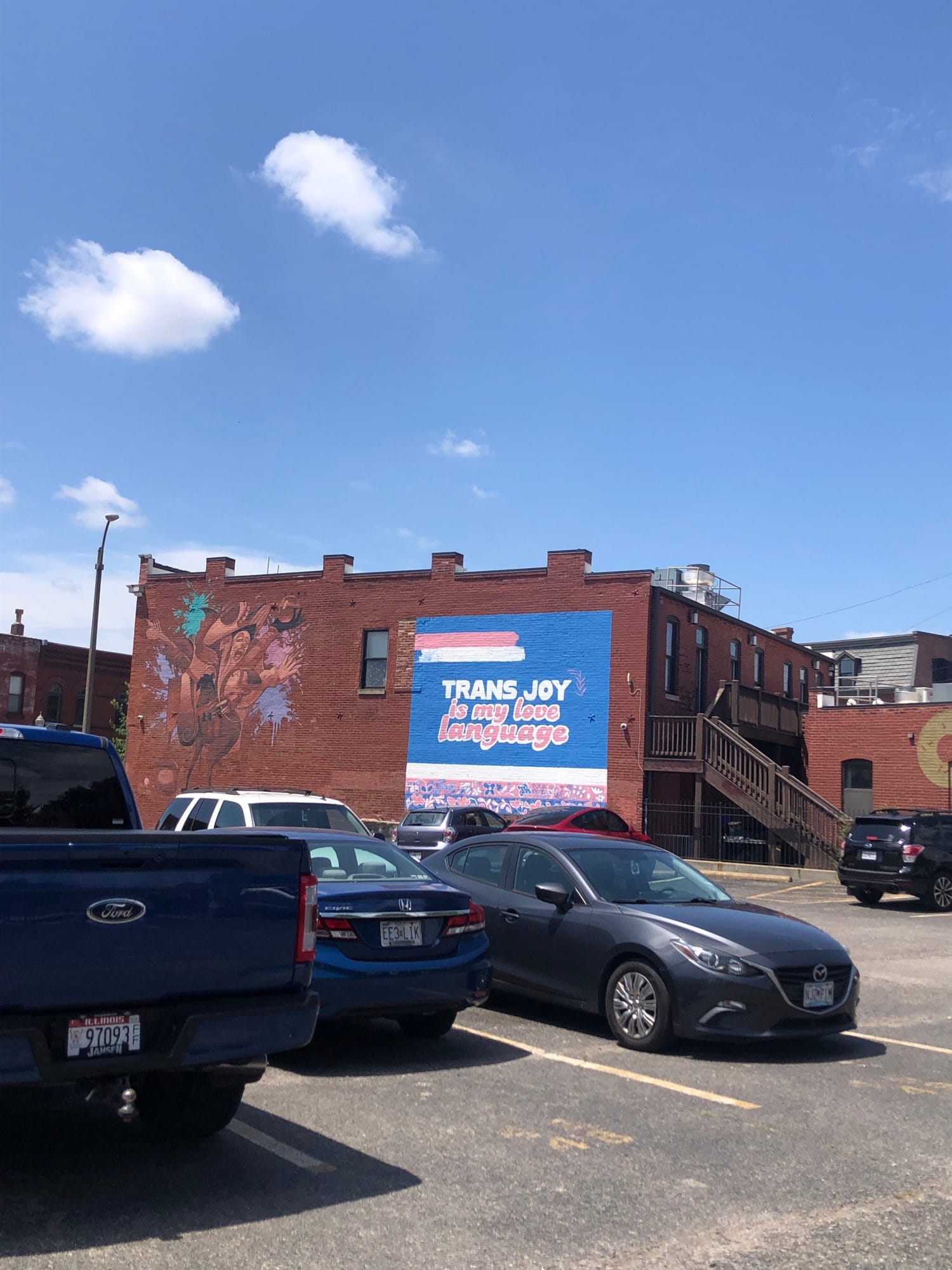 photo of a mural on the side of brick building at the end of a parking lot saying TRANS JOY IS MY LOVE LANGUAGE