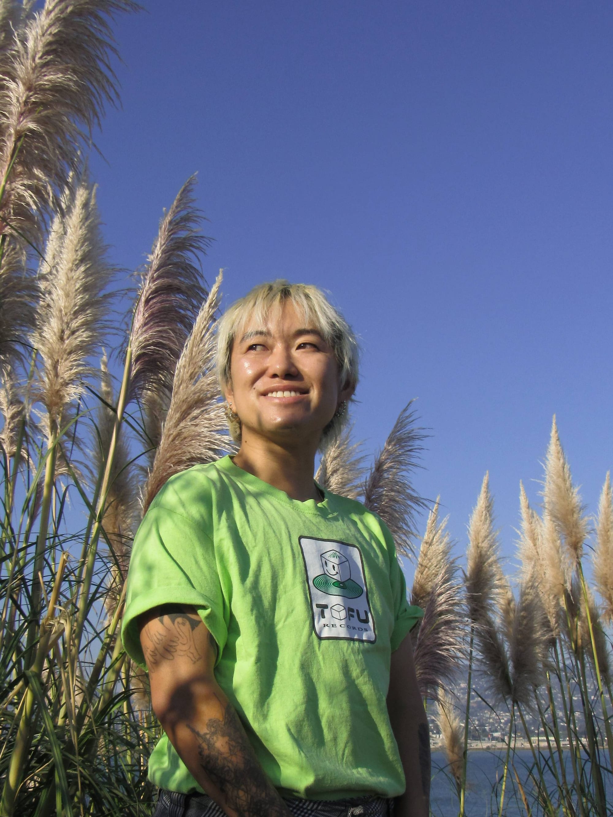 photo of Lio Min wearing a light green shirt with a Tofu Records logo, standing in front of a grove of fluffy pampas grass