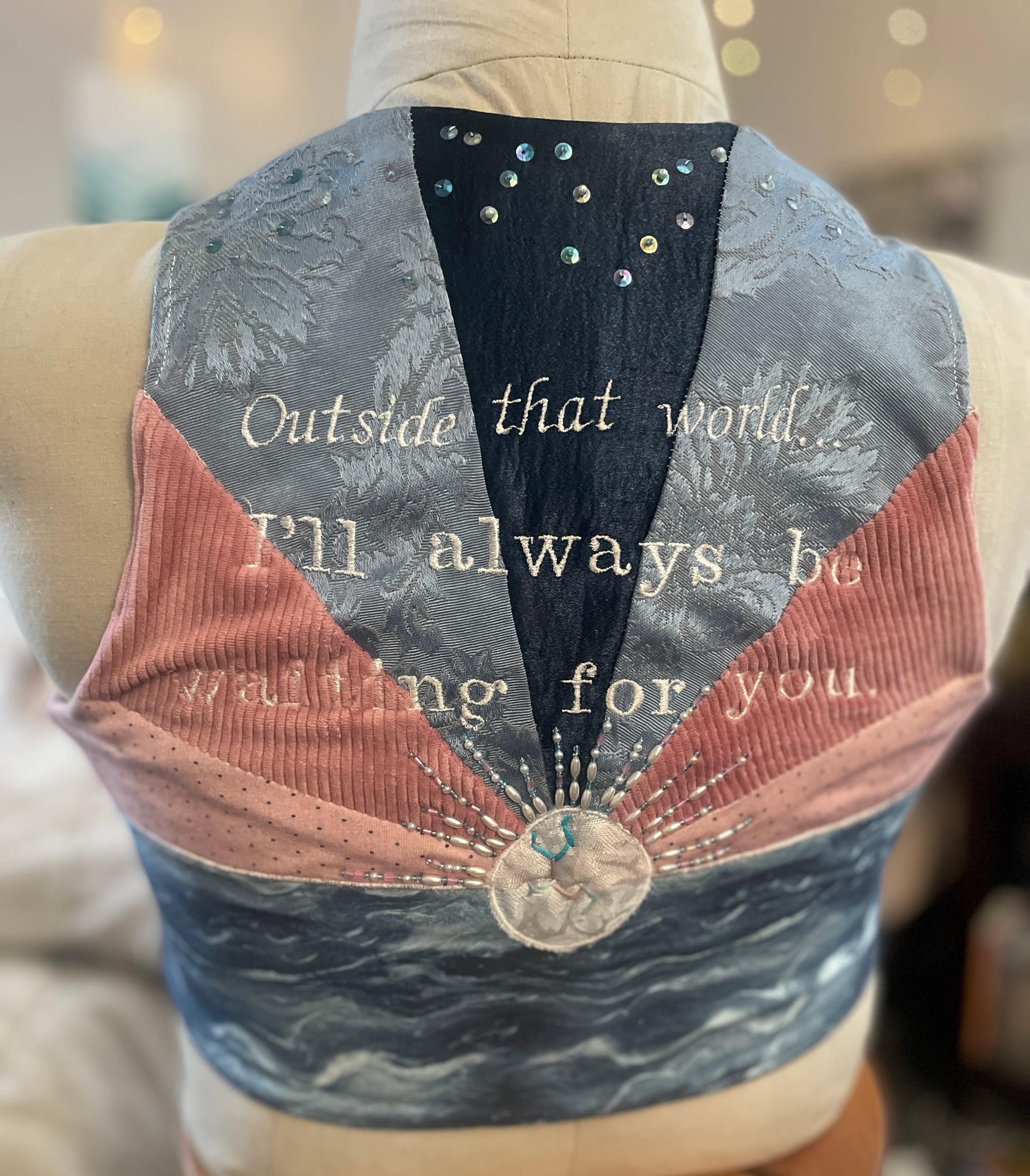 A binder with the trans pride colors and the words “Outside that world I’ll always be waiting for you” on the back. The binder is displayed on a fabric mannequin and features hand embroidered beading and sequins. 