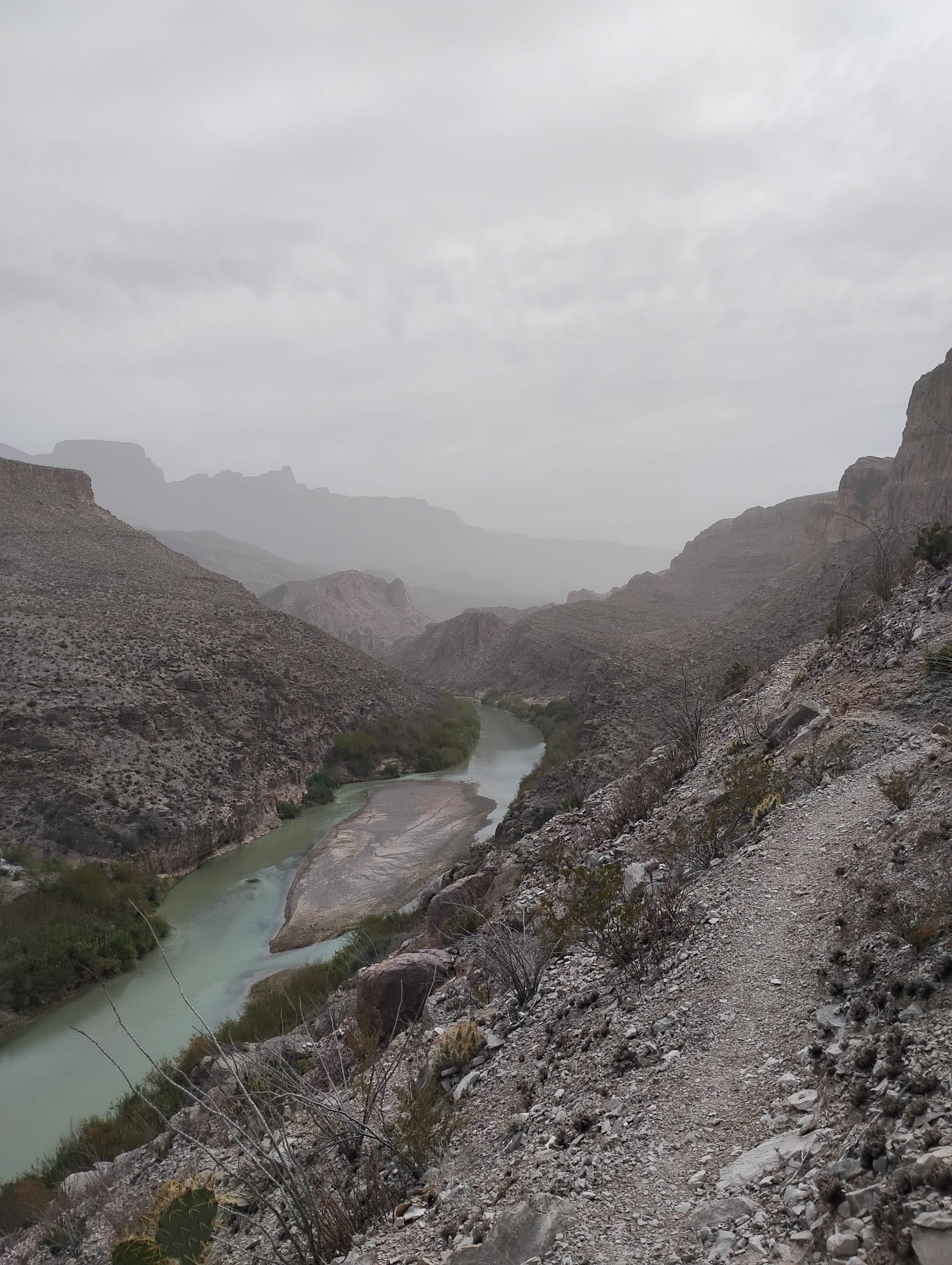 A foggy desert canyon with a winding green river in the bottom, leading off to towering, rugged mountains. A thin footpath winds along the side of the precipitous slope ahead.