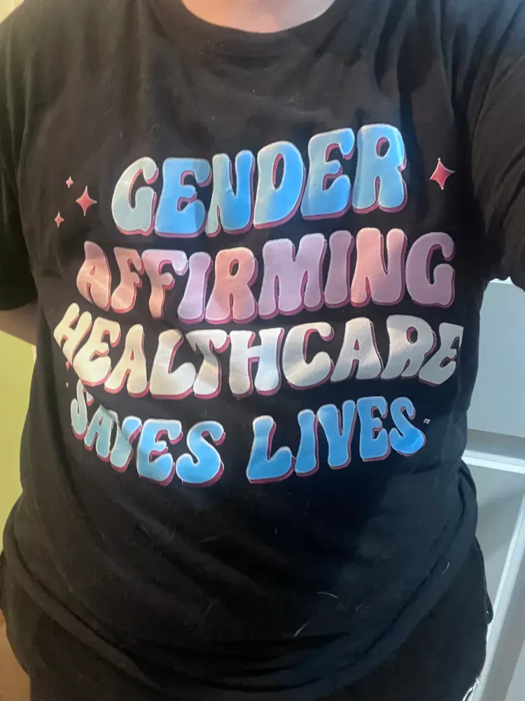 Black t-shirt that says Gender Affirming Healthcare Save Lives in pink, white, and blue bubble letters, with pink accent stars.