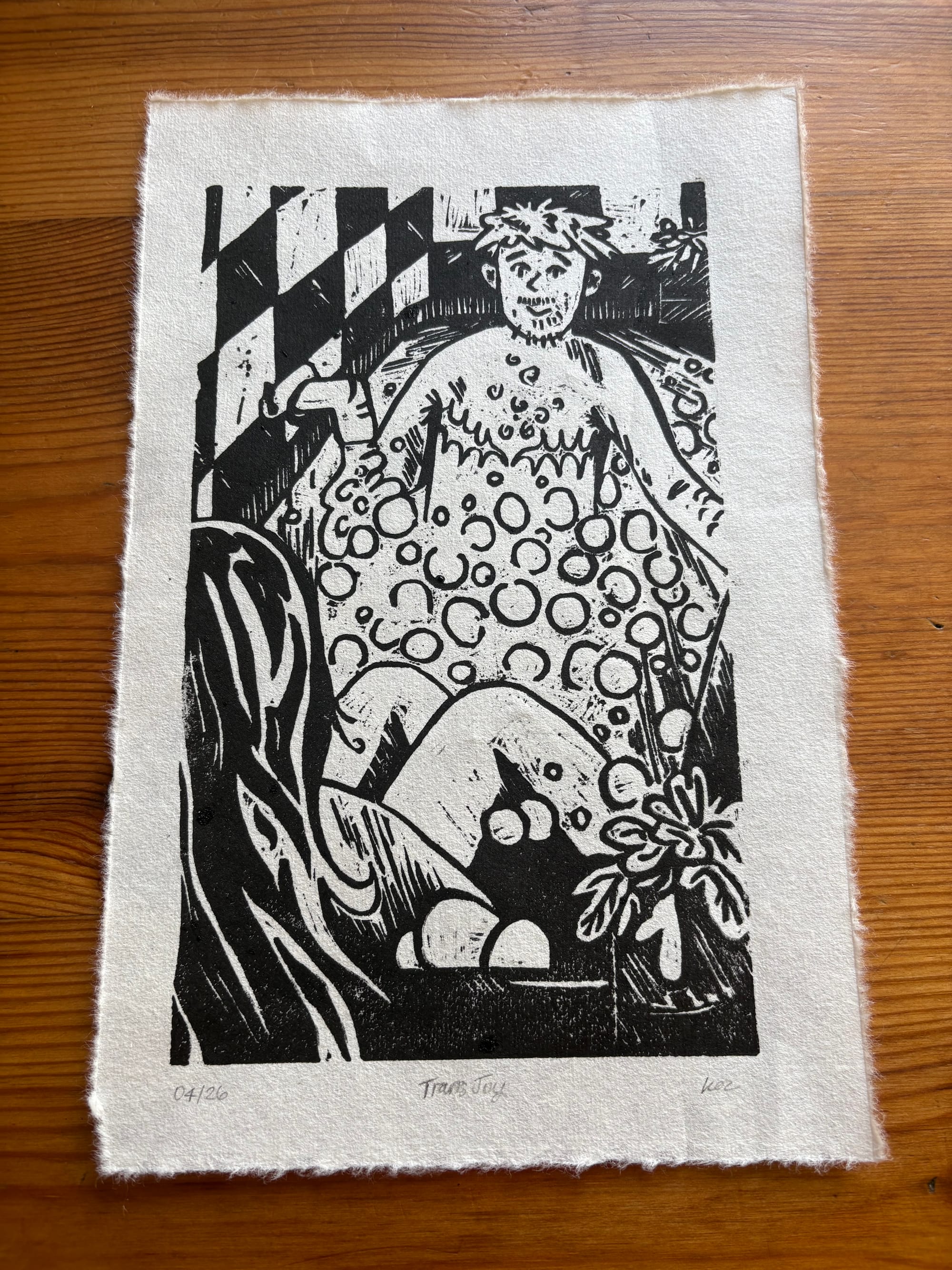 Photograph of a lino cut relief print in black and white. It shows a transmasc figure with stylised top surgery scars reclining in a bathtub. The bubbles are also very stylised. In the background are black and white tiles and a houseplant. The figure is smiling towards the viewer. They have a very bad moustache and spiky hair. 