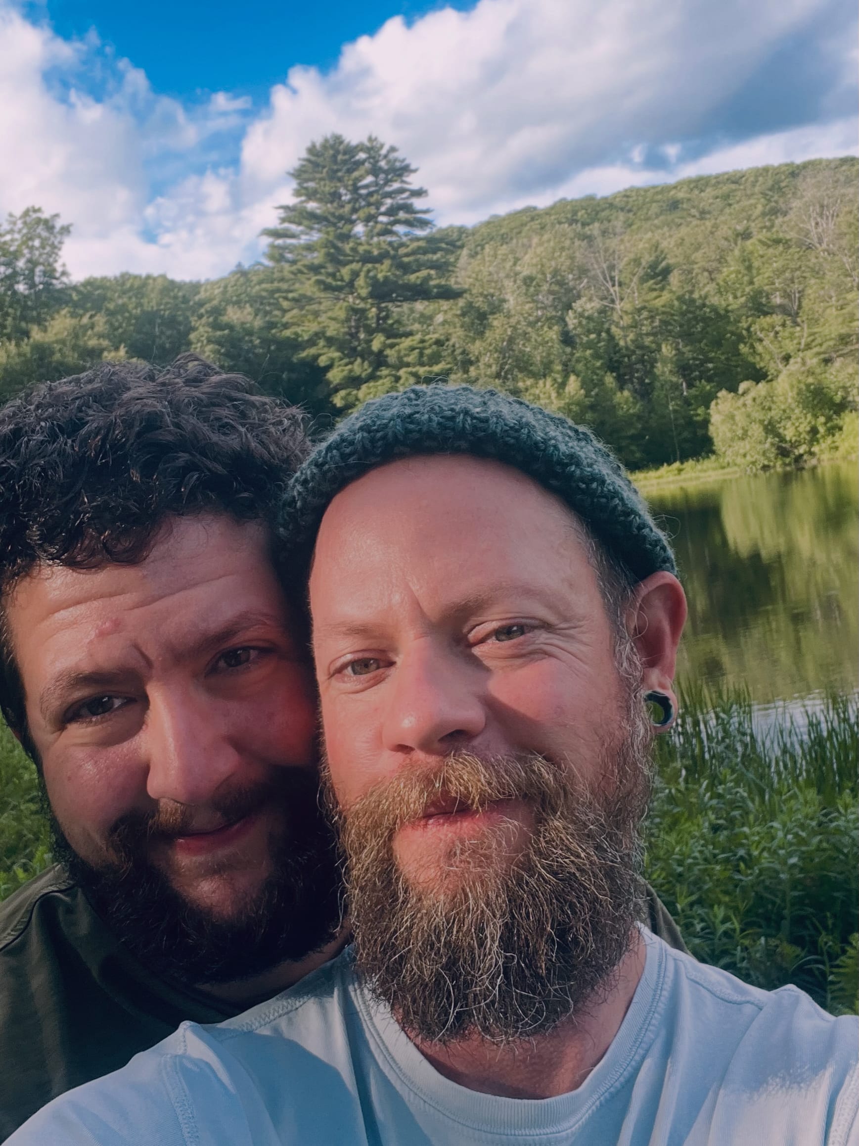 Riley and his husband standing against a beautiful backdrop of blue sky, trees, and water.