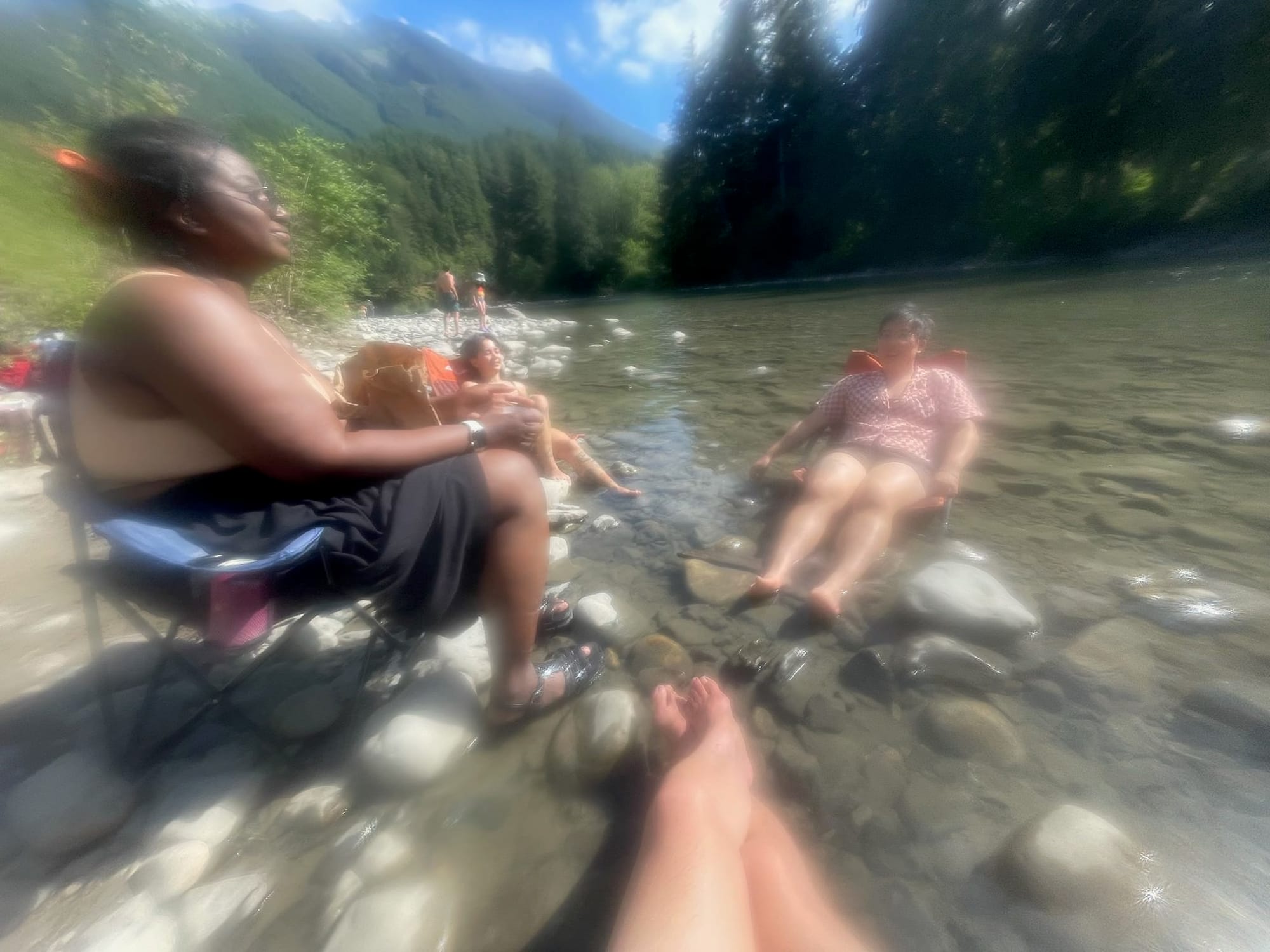 Sunny day with three trans friends of color sitting in the river shallows on camping chairs. Green mountain and blue sky in the background.