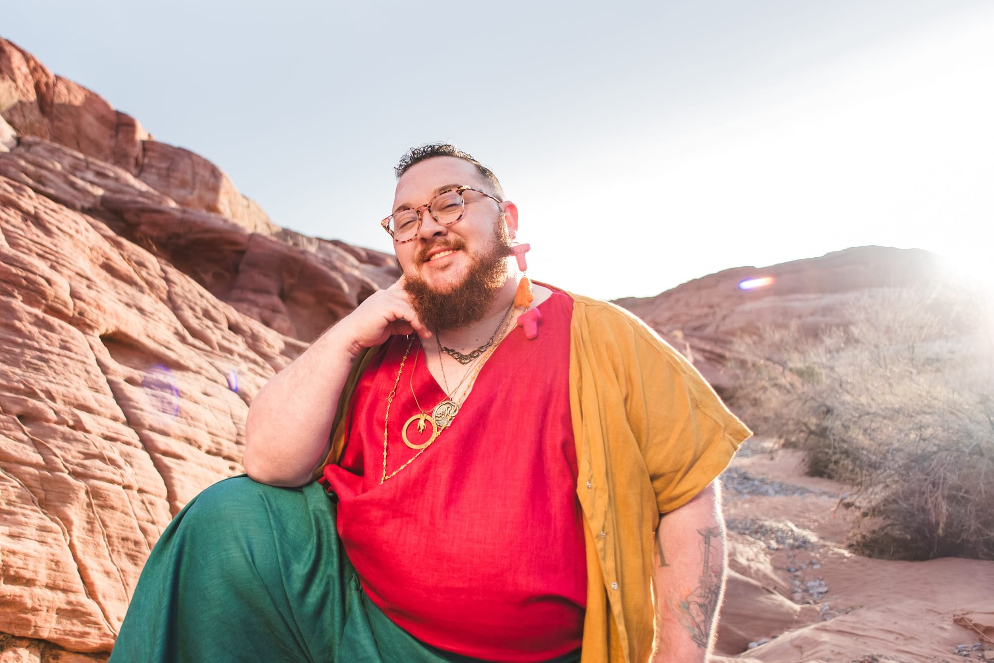 Eli, a white queer and trans person, basks in the sun before Arizona rocks, in a colorful outfit of yellow, red, and green.