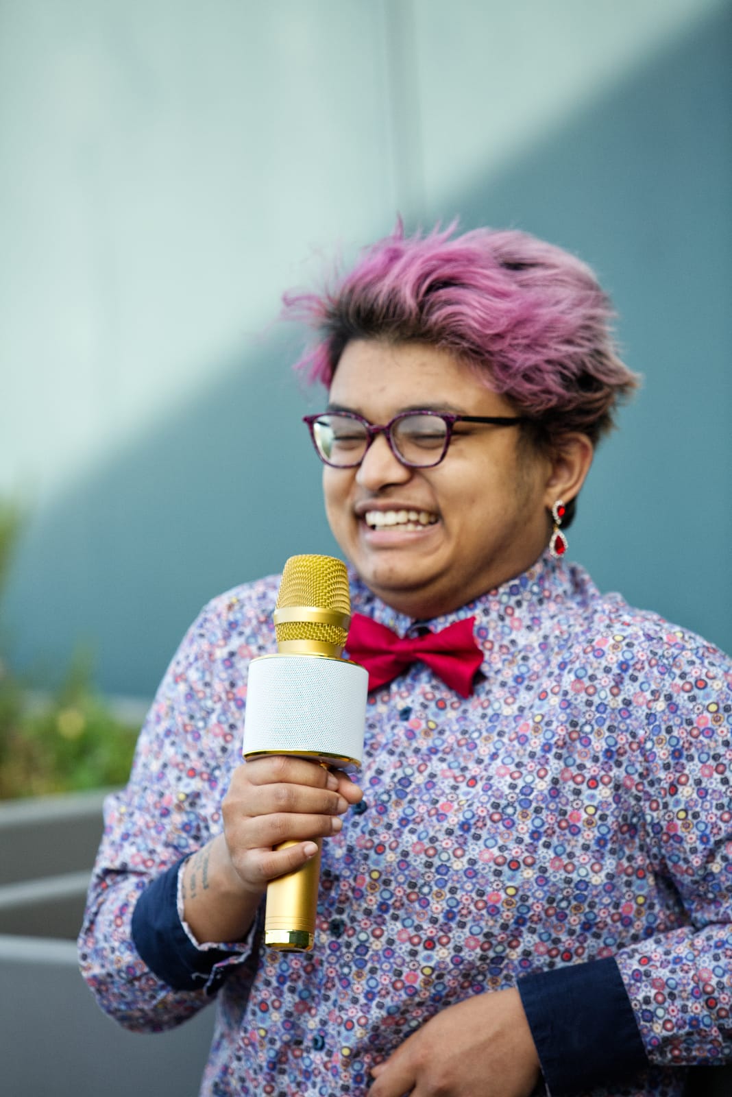 Yoshi, a Brown trans boy with pink hair, wears a patterned shirt and red bow tie, holding a gold microphone and smiling.