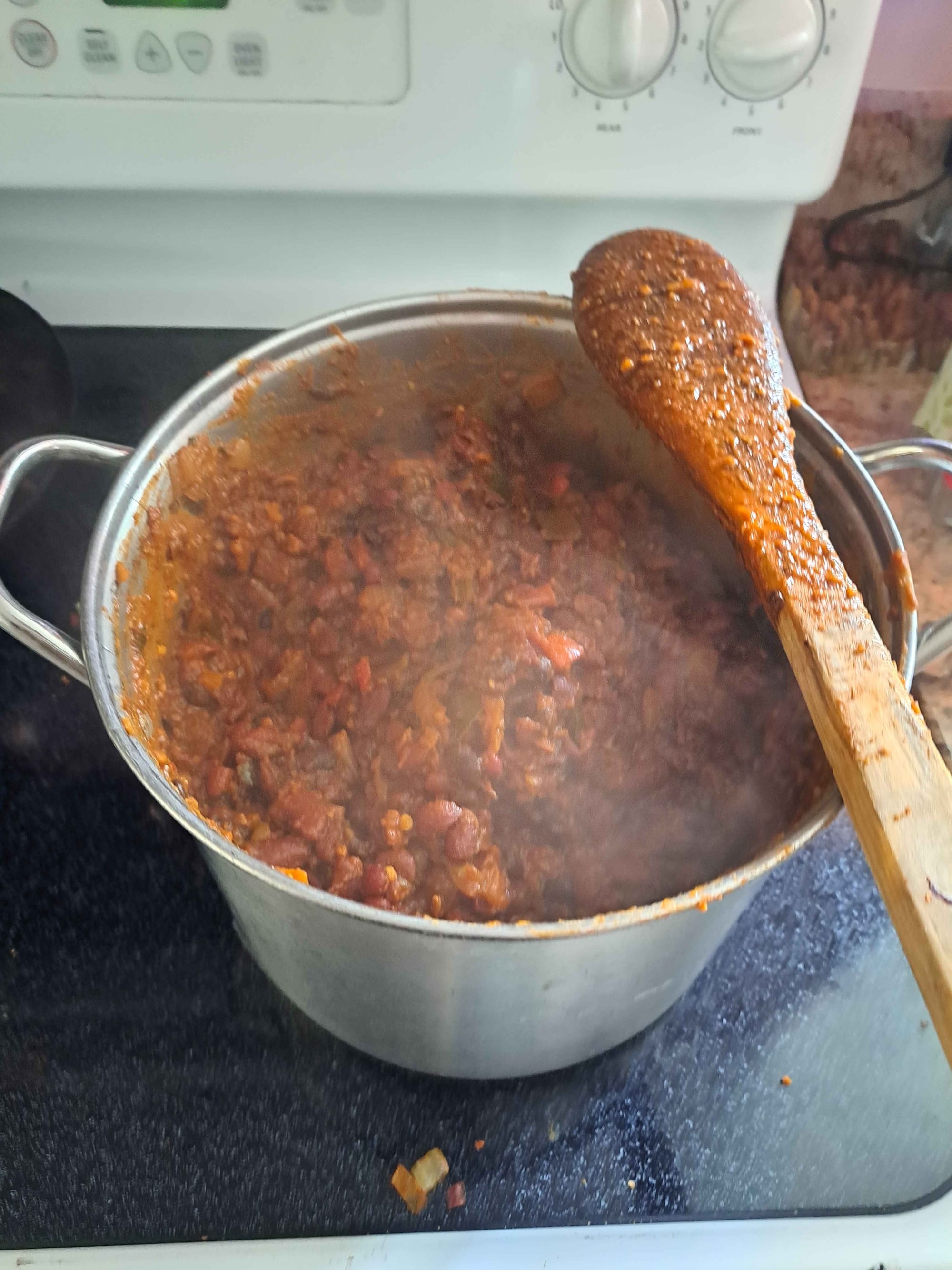A large aluminum stockpot sits on a black stovetop, filled with steaming red chili. A wooden spoon rests across the top, coated in sauce. Steam rises from the surface, and bits of onion and beans are visible in the thick mixture.