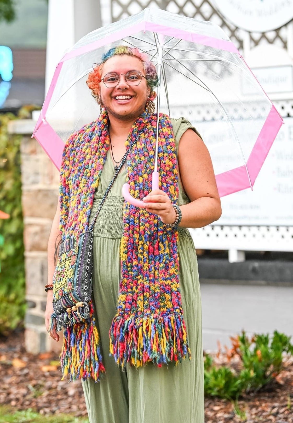 Marco stands outdoors smiling broadly, holding a clear umbrella with pink trim. They have short multicolored hair dyed in sections of blue, orange, and pink, and wear glasses, an olive-green jumpsuit, and a long, vibrant crocheted scarf. Behind them is a building with a white lattice wall.