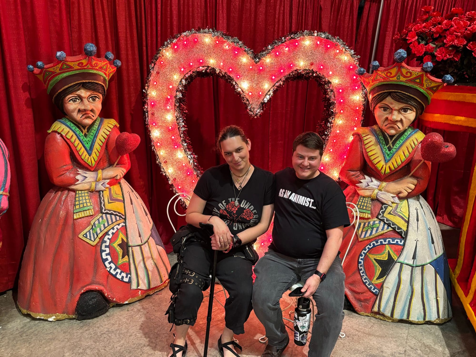 Two people sit close together smiling in front of a large glowing heart-shaped marquee light decorated with pink bulbs. They are flanked by two oversized painted figures of crowned queens in ornate, brightly colored dresses, each holding a glittery red heart, set against a deep red curtain backdrop. Both wear black t-shirts; one has a cane resting beside them.