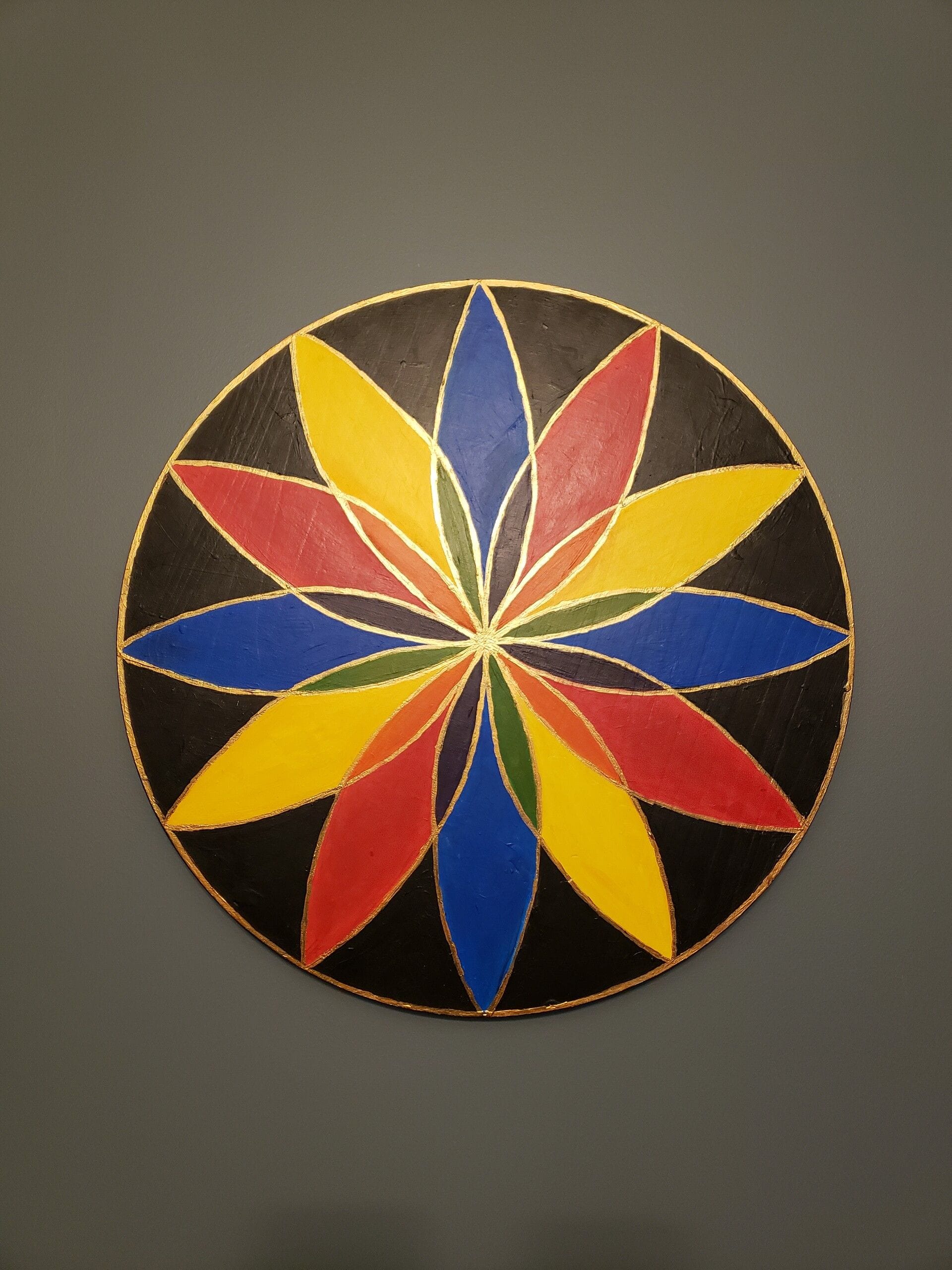 A circular wooden plaque painted in the folk art tradition of Pennsylvania German barn hexes. The design is a twelve-pointed rosette made of overlapping petals alternating between primary colors — red, yellow, and blue — against a black background. Where the petals overlap, they create secondary colors: orange where red meets yellow, green where yellow meets blue, and purple where blue meets red. Thin gold outlines trace each petal and the outer edge of the circle, giving the piece a stained-glass effect. The wood grain is faintly visible beneath the paint, and the plaque hangs against a neutral gray wall.