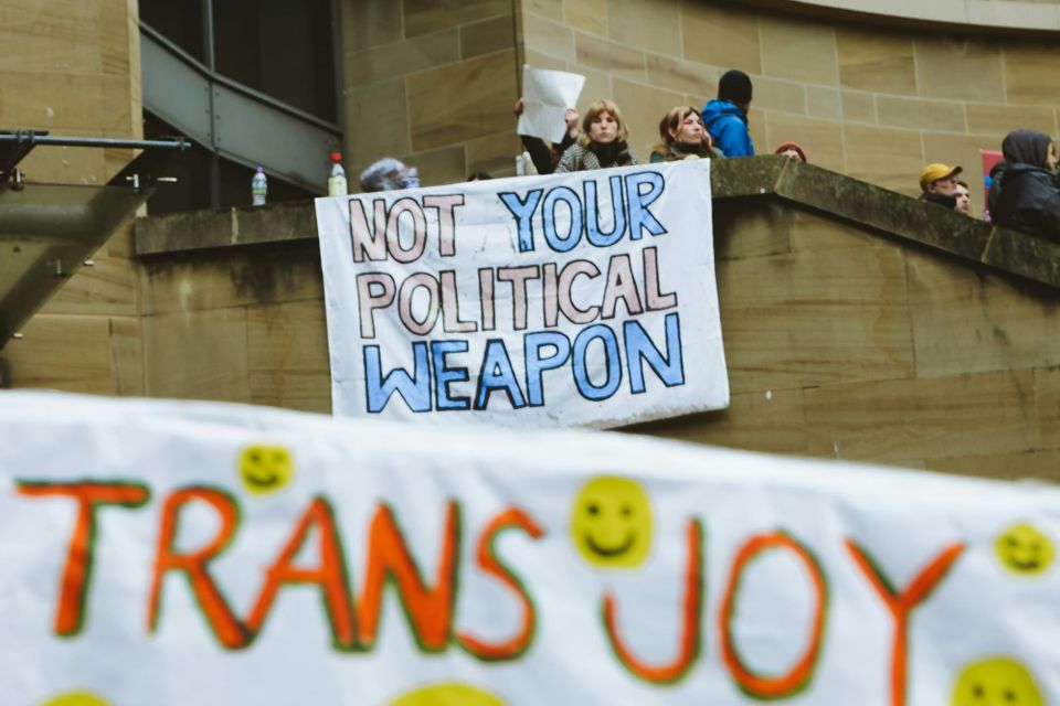 a sign in trans colors that says Not your political weapon in the background, and in front, a banner with smiley faces that says Trans Joy in all capital orange letters.