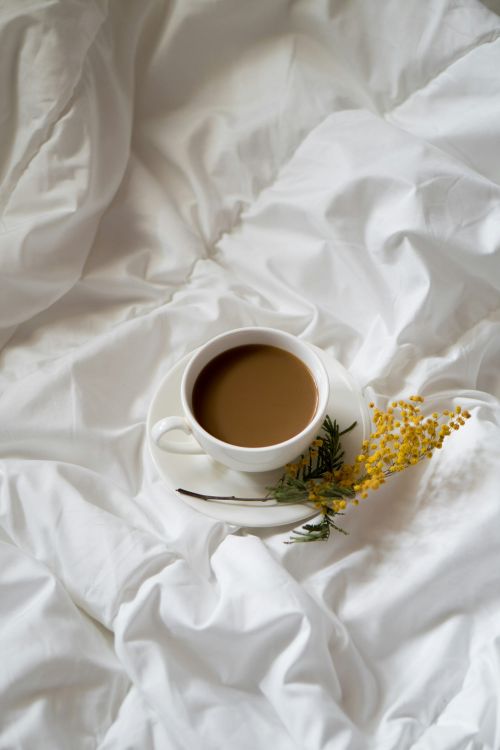 A cup of coffee on a bed with a yellow flower.