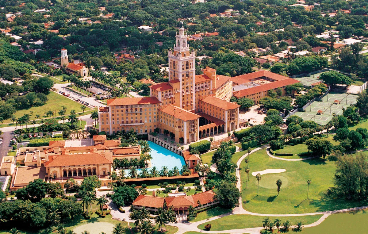 Aerial view of the Biltmore Hotel, a boutique luxury hotel in Coral Gables, showcasing its Mediterranean Revival architecture, red-tiled roof, and historic swimming pool surrounded by tropical palm trees