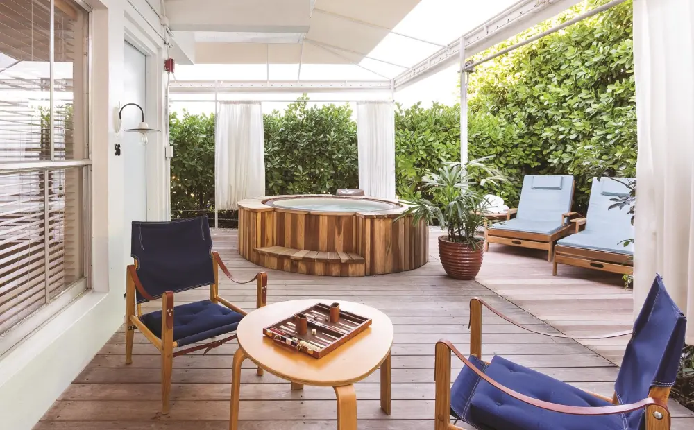 Private terrace at The Standard Spa, Miami Beach with wooden hot tub, blue lounge chairs, white curtains, and a small table set for backgammon.
