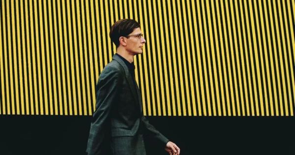 Official portrait of Mark Adam Harold walking in front of a wall with painted black and yellow stripes, photo by Arcana Femin