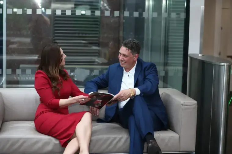 Elliott King in a navy suit and Aleksandra King in a red dress share a magazine on an office sofa.