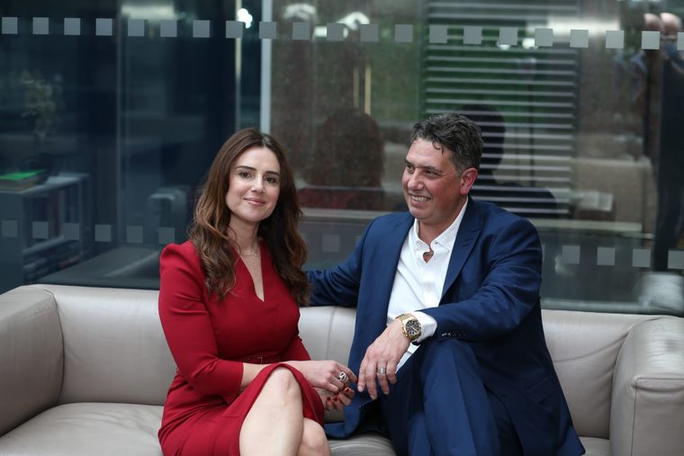 Elliott King rests an arm on the sofa beside Aleksandra King, both smiling in a glass office lobby.