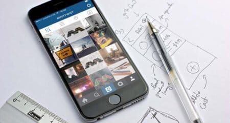 An iPhone showing an Instagram profile grid rests on a desk beside a hand-drawn layout sketch and pen.